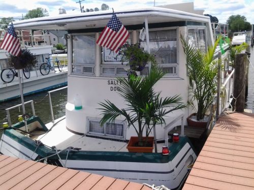A decorated houseboat with American flags and potted plants, docked at a marina on a sunny day.