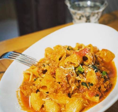 A plate of pasta with meat sauce, garnished with herbs, served with a fork and a glass of water in the background.