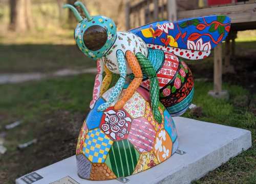 Colorful bee sculpture with intricate patterns and designs, sitting on a stone base in a grassy area.