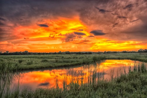 A vibrant sunset over a tranquil river, with colorful clouds reflecting on the water and lush greenery surrounding the scene.
