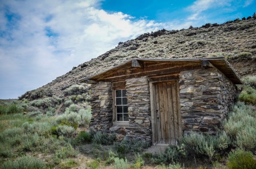 A rustic stone cabin nestled in a grassy landscape, surrounded by hills under a partly cloudy sky.