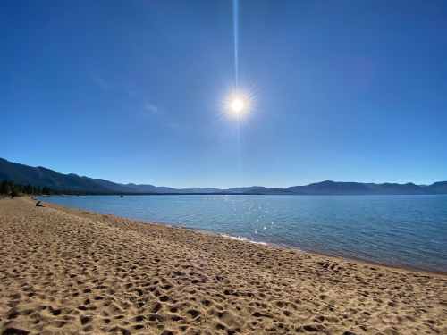 A serene beach scene with golden sand, calm water, and a bright sun shining over distant mountains.