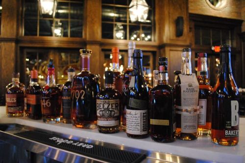 A variety of whiskey bottles displayed on a bar counter in a warm, rustic setting.