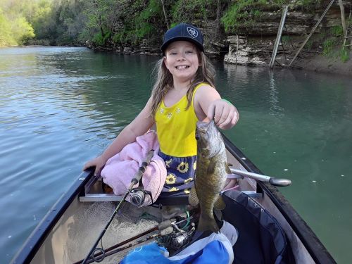 A smiling girl in a yellow tank top holds a fish while sitting in a canoe on a calm river surrounded by greenery.