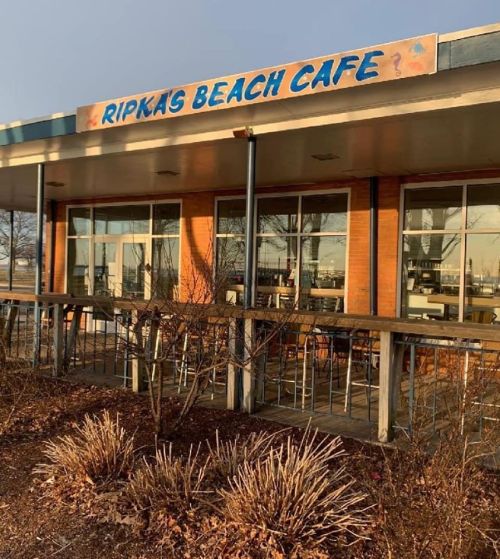 Exterior of Ripkas Beach Cafe, featuring a sign and large windows, with a view of a patio and surrounding plants.
