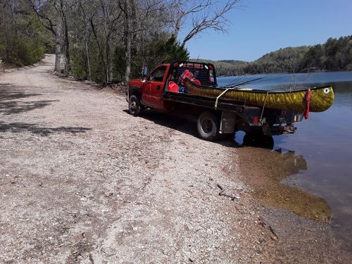 A red pickup truck parked near a calm lake, with a kayak secured in the truck bed and trees in the background.