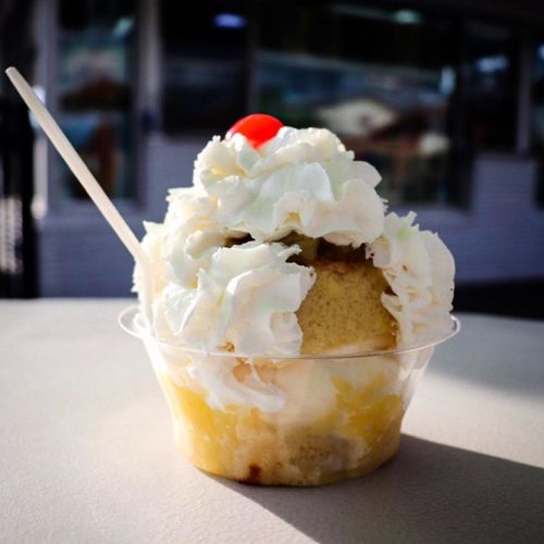 A dessert cup with layers of cake, whipped cream, and a cherry on top, set against a blurred background.