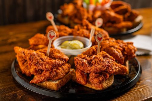 A platter of crispy fried chicken pieces on bread, served with a small bowl of sauce and garnished with toothpicks.