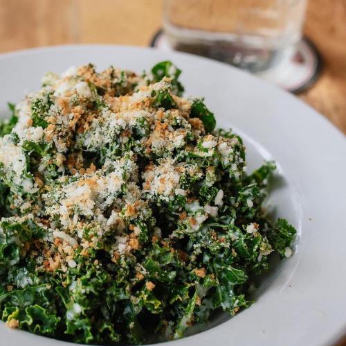 A close-up of a fresh kale salad topped with breadcrumbs, served in a white bowl on a wooden table.