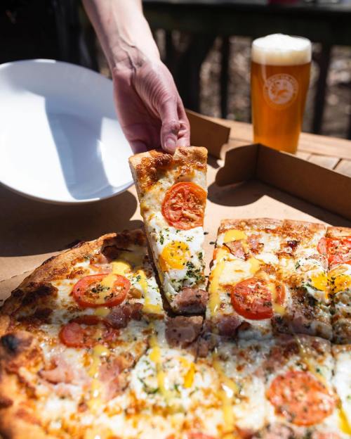 A hand lifts a slice of pizza topped with tomatoes and cheese from a box, with a beer and empty bowl nearby.