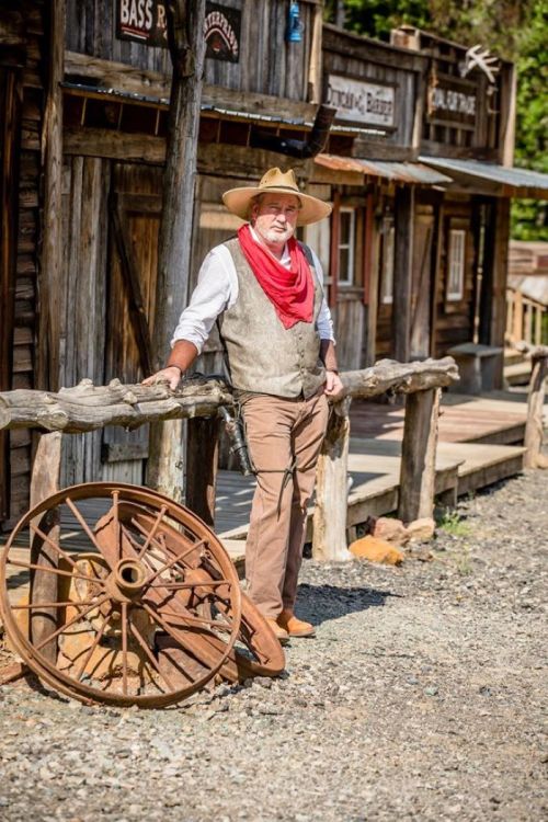 A man in a cowboy hat and red bandana stands by a wooden fence near an old wagon wheel in a rustic western town.
