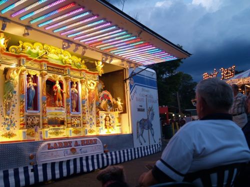A person sits in front of a colorful carnival attraction with lights, watching a performance as dark clouds gather above.