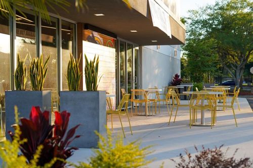 Outdoor seating area with yellow chairs and tables, surrounded by potted plants and greenery.