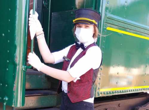 A young train conductor in a mask and gloves stands by a green train, holding the door open with a welcoming smile.