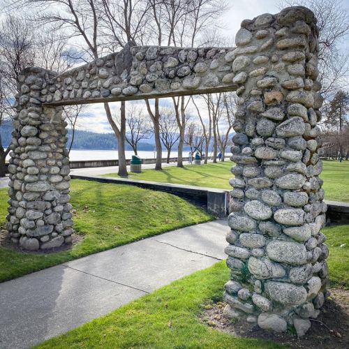 Stone archway in a park, surrounded by grass and trees, with a lake visible in the background under a cloudy sky.