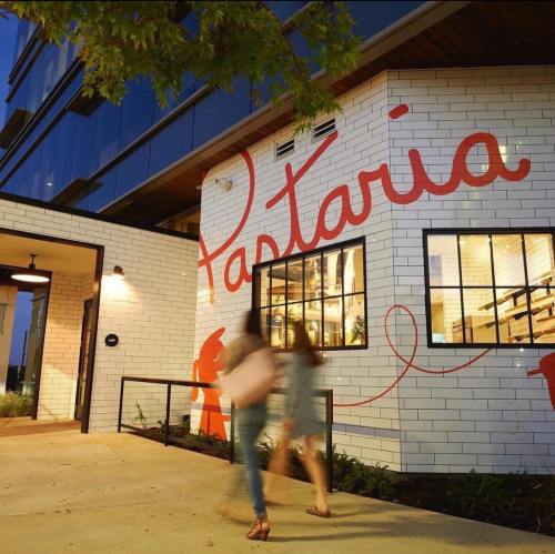 Two people walk past a modern building with a large "Pastaria" mural on the wall, illuminated at dusk.
