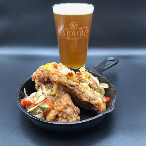 A cast iron skillet filled with crispy fried chicken and colorful vegetables, alongside a glass of beer.