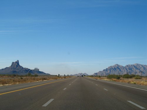 A wide highway stretches into the distance, flanked by mountains under a clear blue sky.