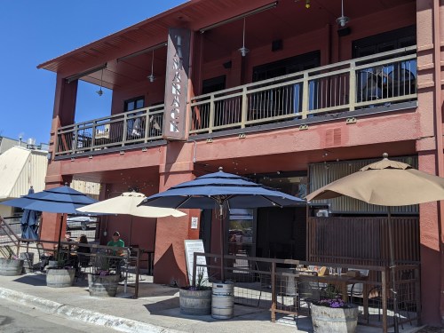 A restaurant exterior with outdoor seating, umbrellas, and a sign reading "Tamarack" under a clear blue sky.
