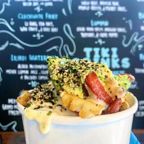A bowl of fries topped with avocado, bacon, sesame seeds, and drizzled with sauce, against a colorful wall backdrop.