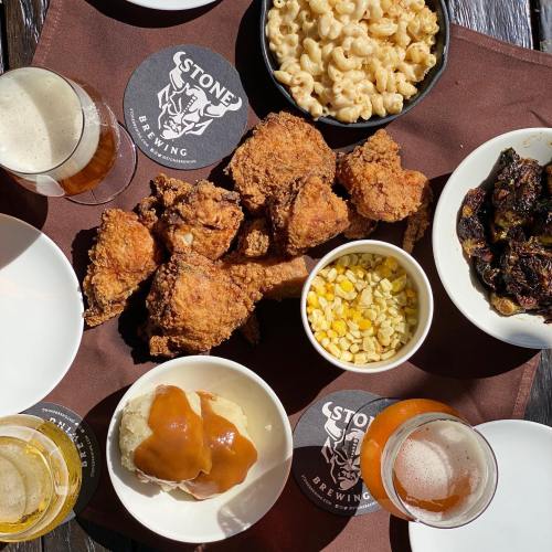 A spread of fried chicken, macaroni and cheese, corn, mashed potatoes with gravy, and craft beers on a wooden table.