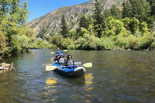 Three people kayaking on a calm river surrounded by lush greenery and mountains under a clear blue sky.