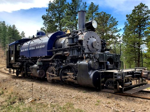 A vintage steam locomotive on a track surrounded by trees and hills under a blue sky.