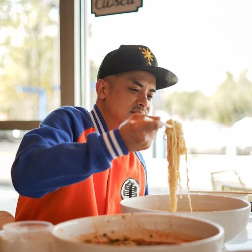 A person in a colorful jacket enjoys a bowl of noodles, focused on lifting noodles with chopsticks.