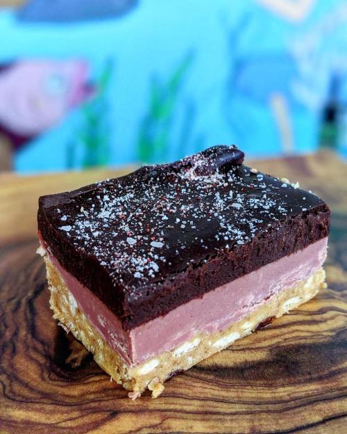 A layered dessert with a chocolate topping, pink filling, and a crumbly base, sprinkled with salt on a wooden surface.