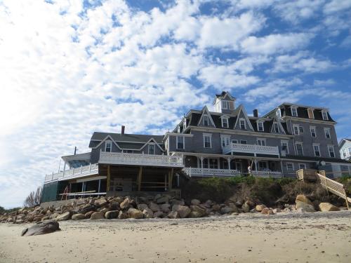 A large, historic beachside hotel with a gray facade, surrounded by rocks and a cloudy blue sky.