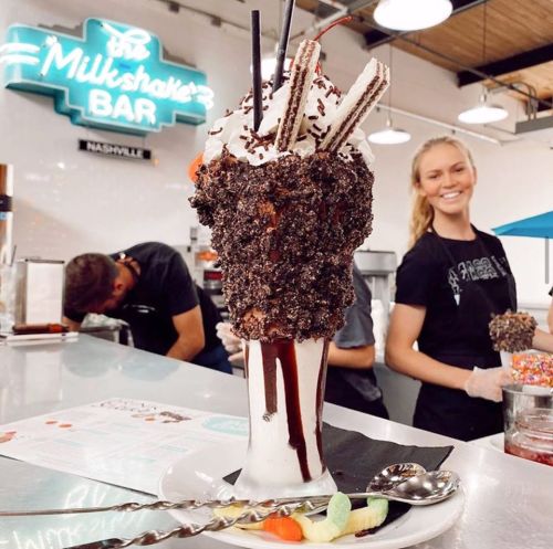 A tall milkshake topped with whipped cream, chocolate, and cookies, with staff preparing in the background at a vibrant bar.