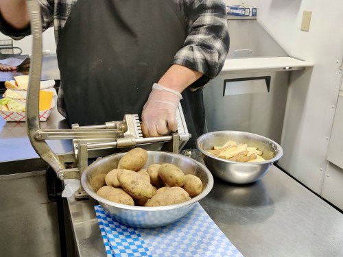 A person using a potato cutter, with a bowl of whole potatoes and a bowl of sliced potatoes nearby.
