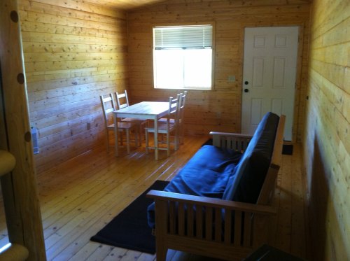 Cozy wooden interior of a cabin featuring a table, chairs, and a futon by the door and window.