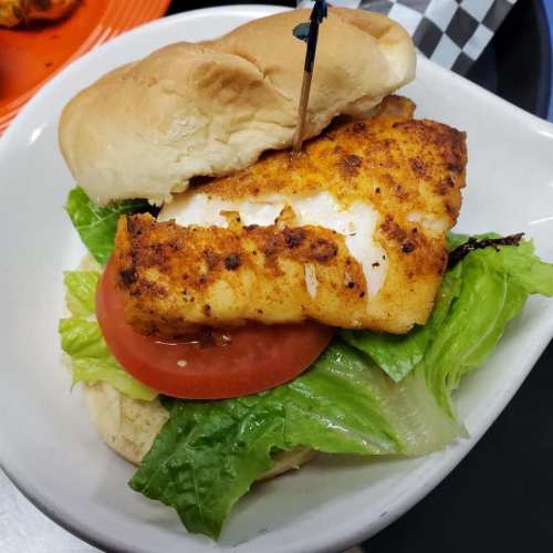 A fish sandwich with seasoned fillet, lettuce, and tomato on a soft bun, held together with a toothpick.