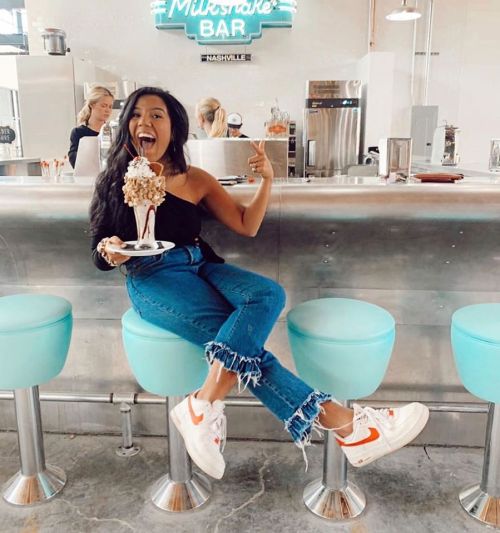 A woman sits on a bar stool, joyfully holding a large milkshake topped with whipped cream and a cherry.