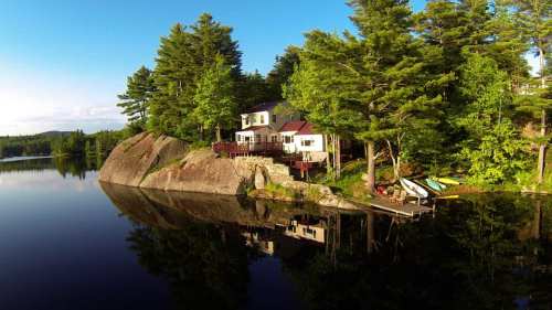 A serene lakeside house surrounded by trees, reflecting in calm water, with kayaks on the shore.