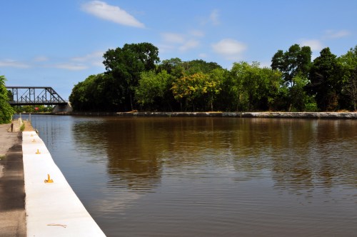 A calm river reflects trees and a blue sky, with a bridge visible in the background along the water's edge.