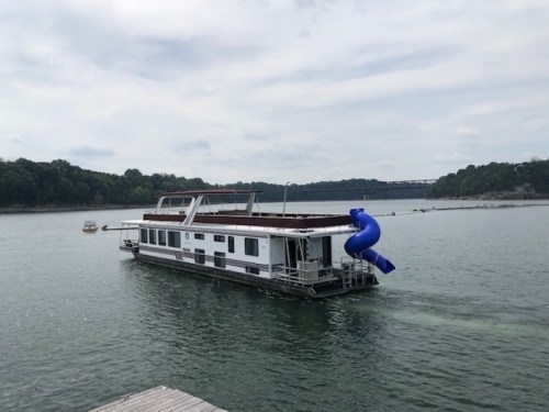 A houseboat with a blue water slide is cruising on a calm lake surrounded by green hills under a cloudy sky.