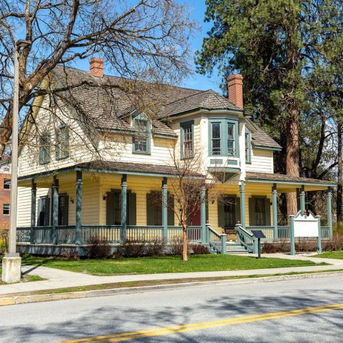 A historic two-story house with a porch, surrounded by trees and a well-maintained lawn on a sunny day.