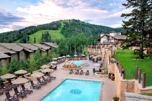A scenic resort with a pool, lounge chairs, and mountains in the background, surrounded by trees and a clear sky.