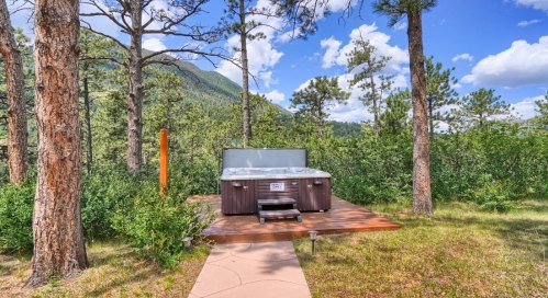A hot tub on a wooden deck surrounded by trees and mountains under a blue sky with fluffy clouds.