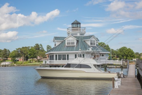 A large white yacht docked beside a charming lighthouse-style house on a calm waterway under a blue sky.