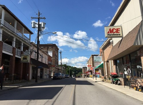A small town street lined with shops, including an Ace Hardware, under a blue sky with fluffy clouds.
