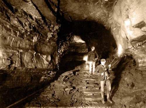 Two miners with torches navigate a rocky underground tunnel, surrounded by dark, textured stone walls.