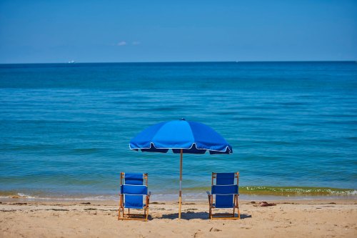 Two blue beach chairs under a blue umbrella on a sandy shore, facing a calm ocean under a clear blue sky.