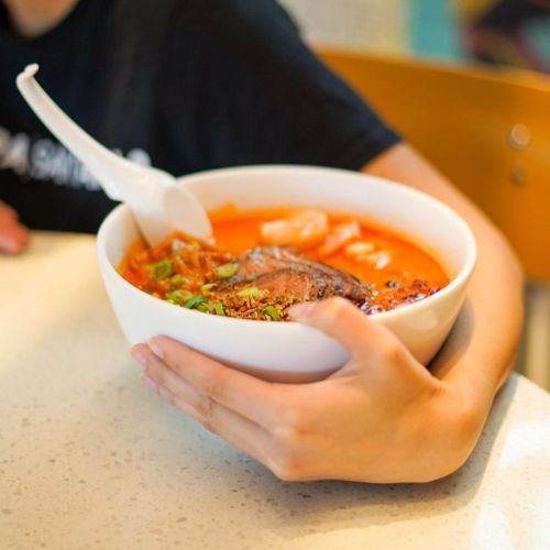 A person holding a large bowl of spicy soup with meat and vegetables, sitting at a table.