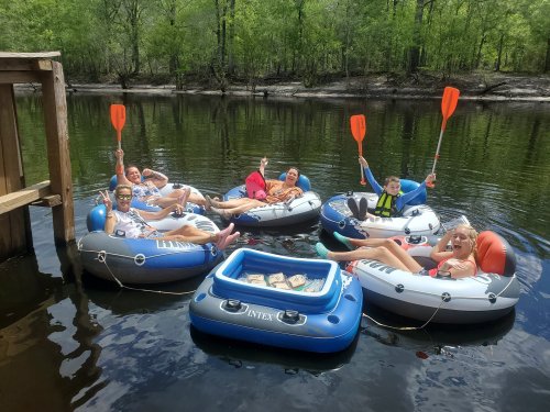 A group of six people in inflatable rafts on a calm river, smiling and holding paddles, surrounded by greenery.