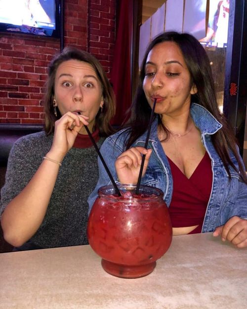 Two women share a large cocktail bowl with straws, smiling and enjoying their drinks in a cozy setting.