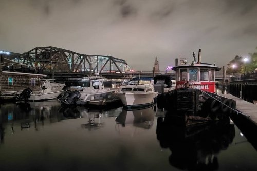 A calm harbor scene at night with boats docked and a bridge in the background under a cloudy sky.