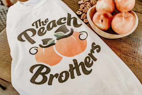 A white t-shirt with "The Peach Brothers" graphic and two peaches, next to a bowl of fresh peaches on a wooden table.